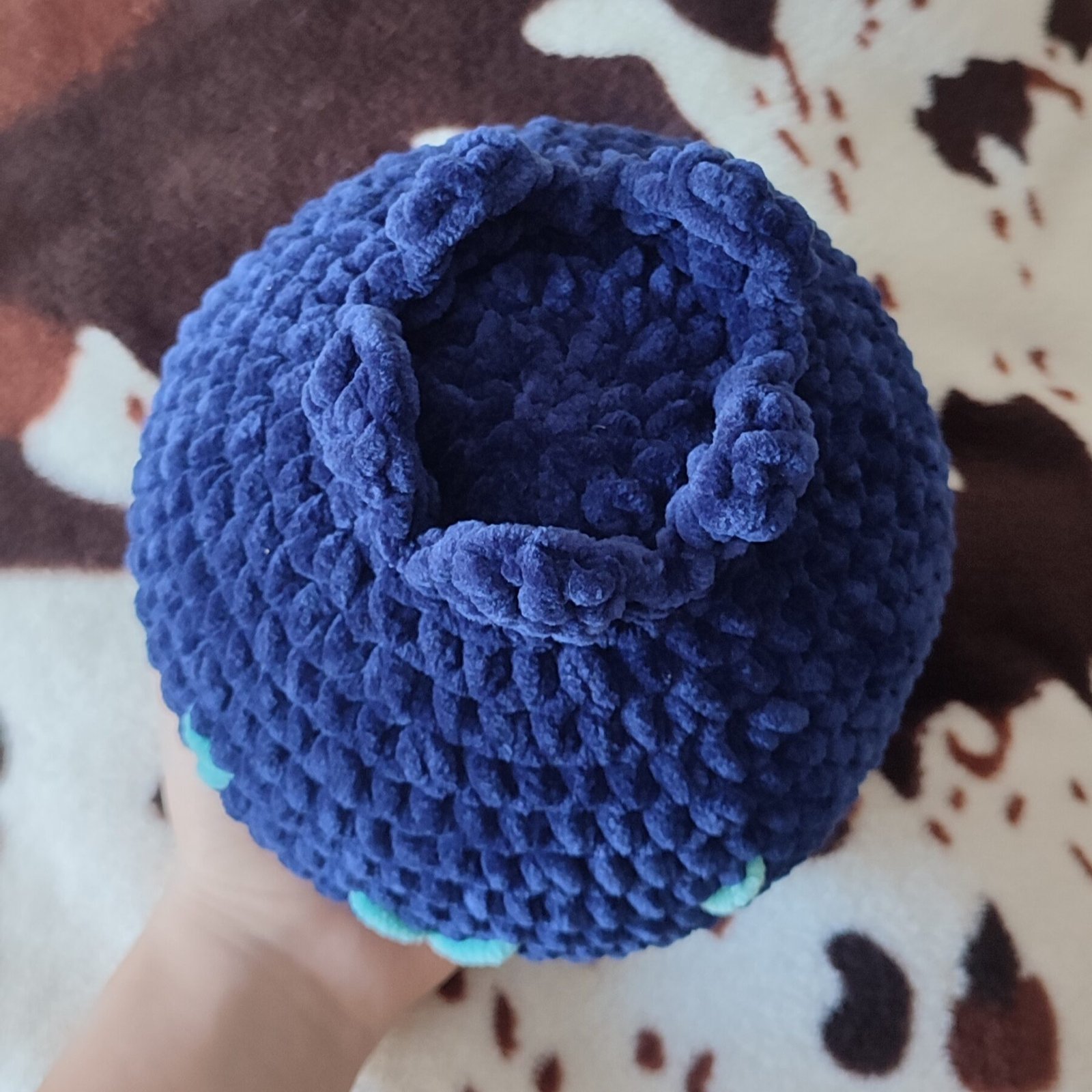 Blueberry plush in a hand, view from top. The photo is taken in a more realistic home setting, in front of a blanket.
