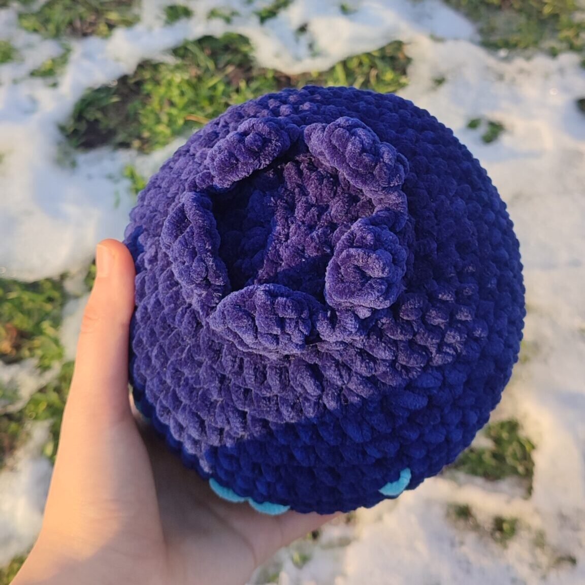 Blueberry plush in a hand, view from top. The photo is taken in front of greenery covered in snow.