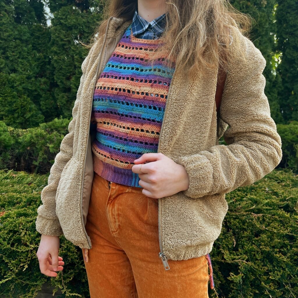 The vest worn on top of a plaid shirt and high-raise jeans. A teddy bear jacket is worn on top of it. The photo is taken outside in front of some greenery.